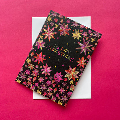 A black Christmas greeting card with colourful star patterns and 'HAPPY CHRISTMAS' text, placed diagonally on top of a white envelope against a bright pink background.