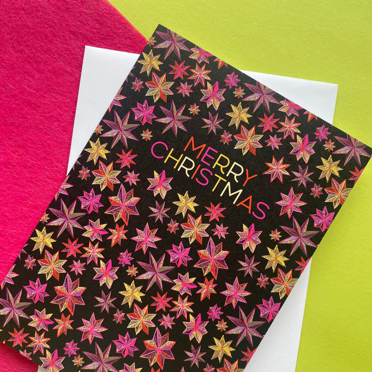 A Christmas greeting card with a black background and a repeating pattern of metallic pink, gold, and orange starbursts, featuring the text 'MERRY CHRISTMAS' in a gold-to-pink gradient. The card is layered over a white envelope, set against a split background of bright pink and lime green.