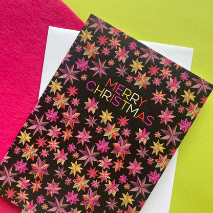A Christmas greeting card with a black background and a repeating pattern of metallic pink, gold, and orange starbursts, featuring the text 'MERRY CHRISTMAS' in a gold-to-pink gradient. The card is layered over a white envelope, set against a split background of bright pink and lime green.