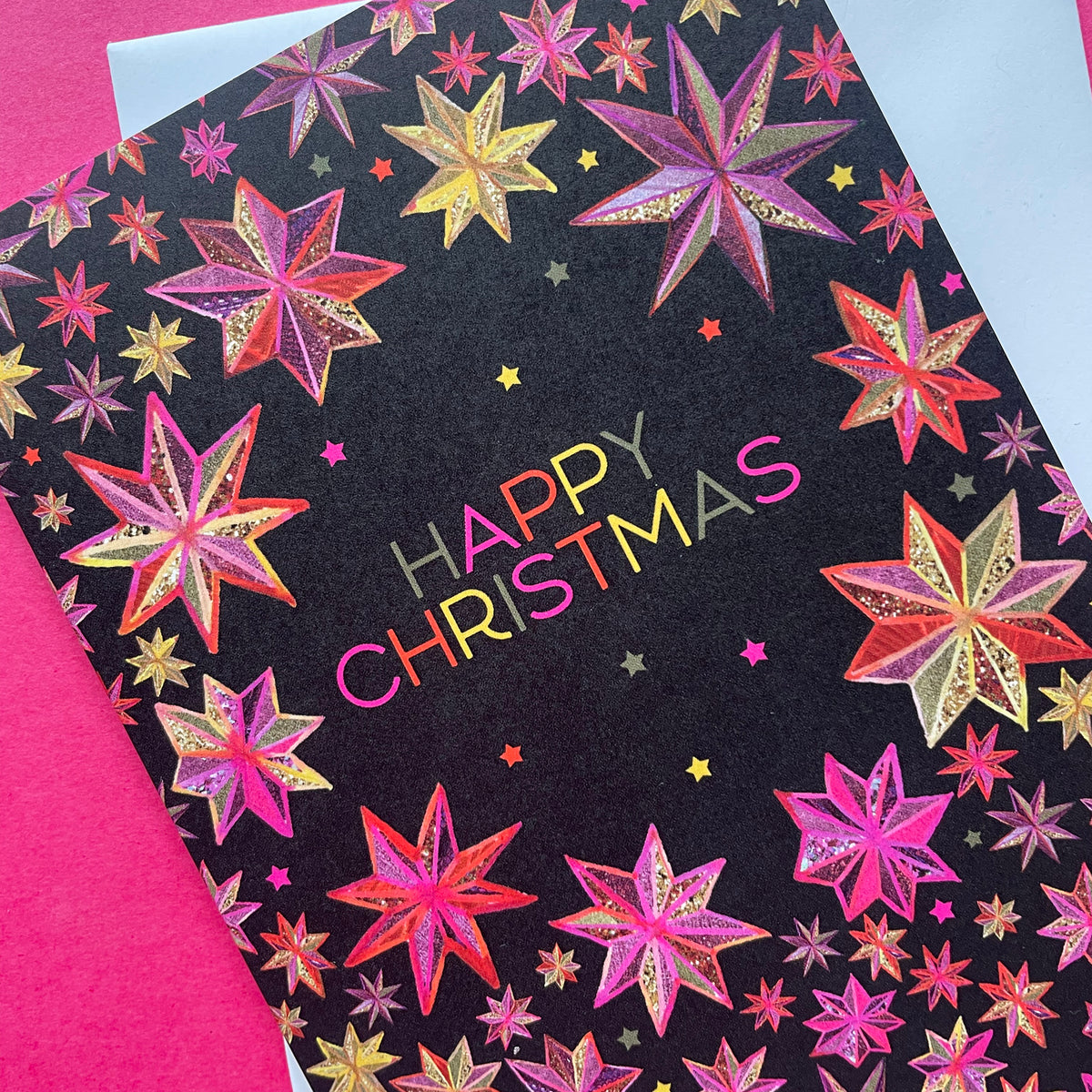 A black Christmas greeting card featuring colourful star patterns and the words 'HAPPY CHRISTMAS' in bright lettering, photographed on a bright pink surface with part of a white envelope visible.