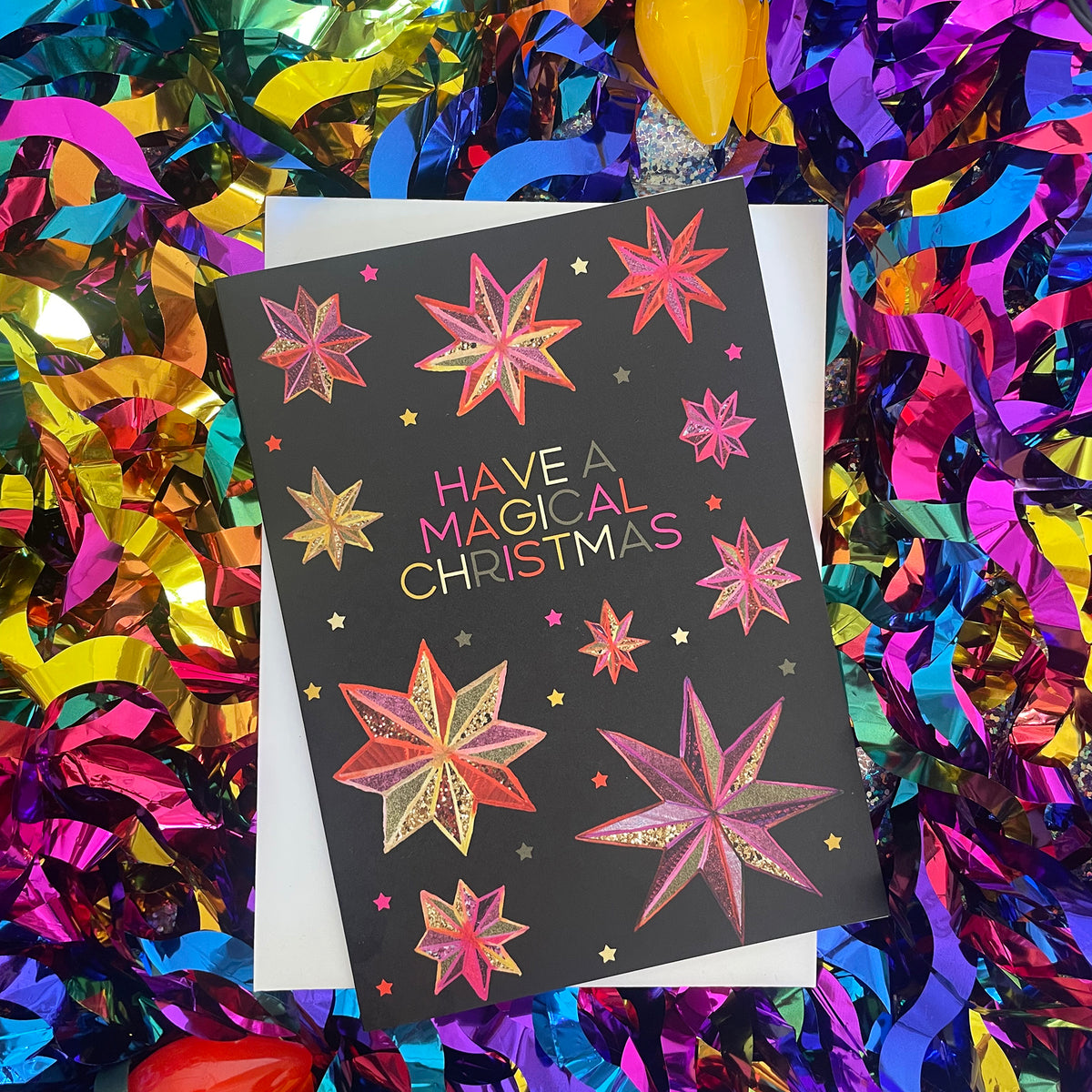 A black Christmas greeting card with hand drawn gold and pink star designs and the text 'HAVE A MAGICAL CHRISTMAS' in gold, pink, and orange lettering, displayed on a white envelope surrounded by colourful metallic tinsel.