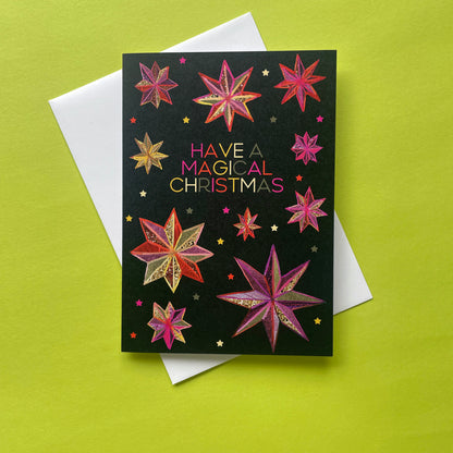 A black Christmas greeting card with colourful star illustrations and the text 'HAVE A MAGICAL CHRISTMAS' in pink, gold, and white lettering, placed on top of a white envelope against a bright lime green background.