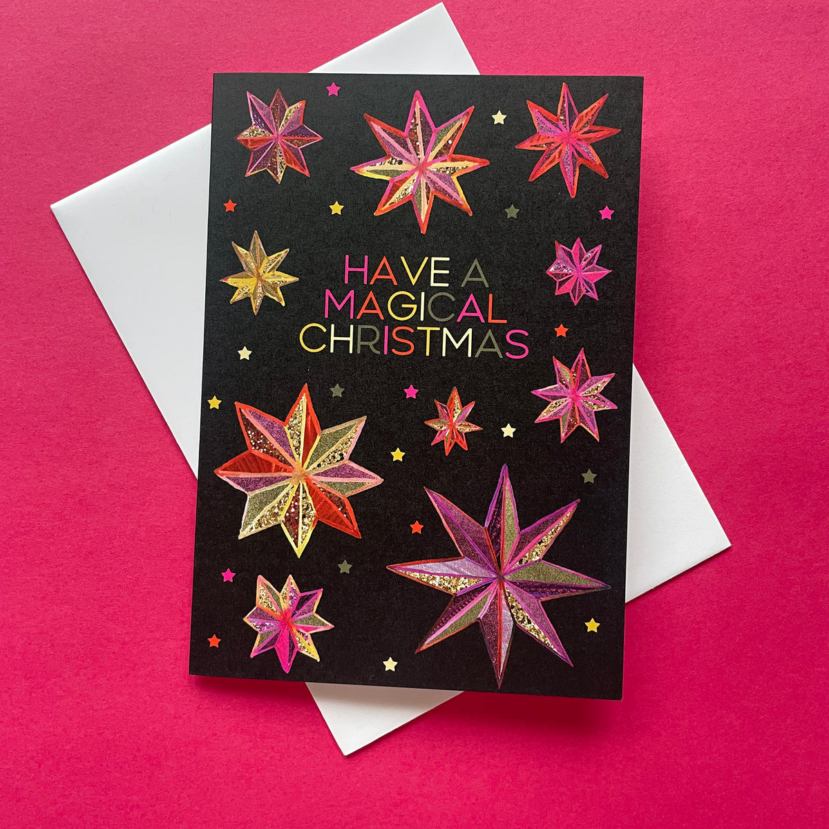 A Christmas greeting card with a black background and colourful stars drawn in pink, gold, and orange, featuring the text 'HAVE A MAGICAL CHRISTMAS' in multicolour lettering. The card is displayed on top of a white envelope against a bright pink background.