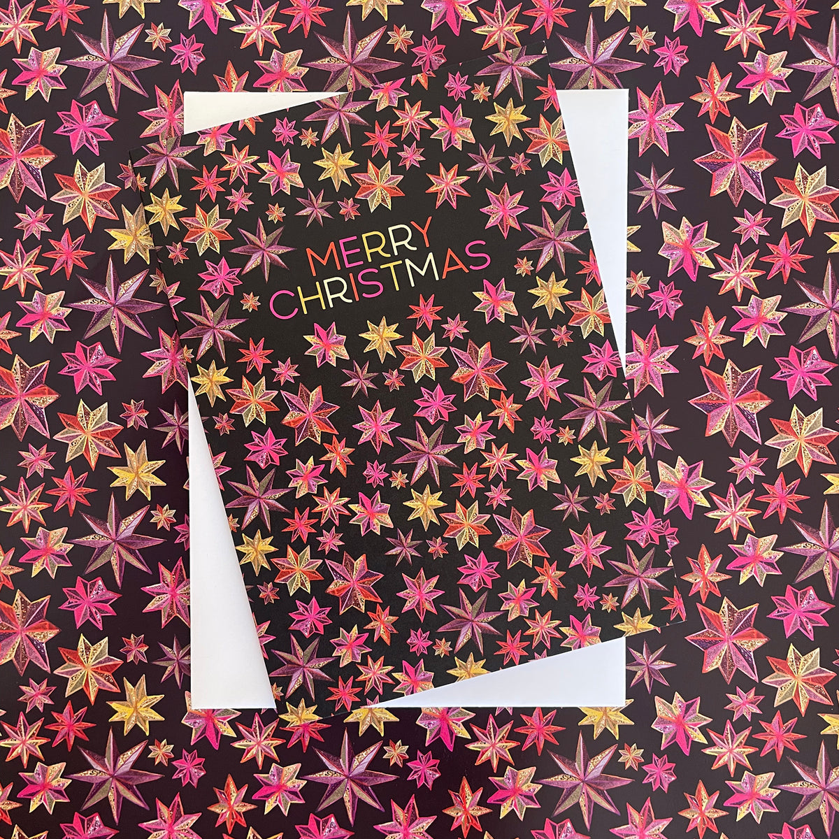 A Christmas greeting card featuring a black background covered in a dense pattern of pink, red, orange, and yellow starburst flowers. The words 'MERRY CHRISTMAS' appear in the center in a multicolour font.