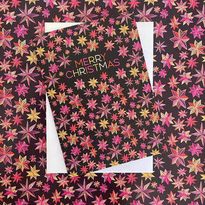 A Christmas greeting card featuring a black background covered in a dense pattern of pink, red, orange, and yellow starburst flowers. The words 'MERRY CHRISTMAS' appear in the center in a multicolour font.