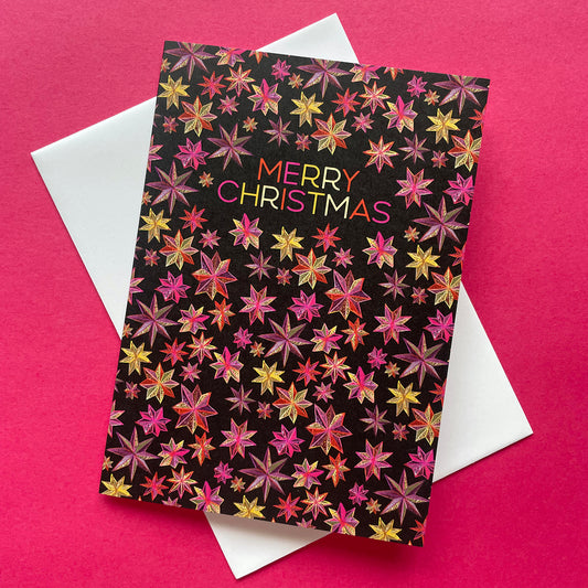 A Christmas greeting card with a black background covered in colourful star patterns and the words 'MERRY CHRISTMAS' in multicoloured lettering, placed on top of a white envelope against a bright pink background.