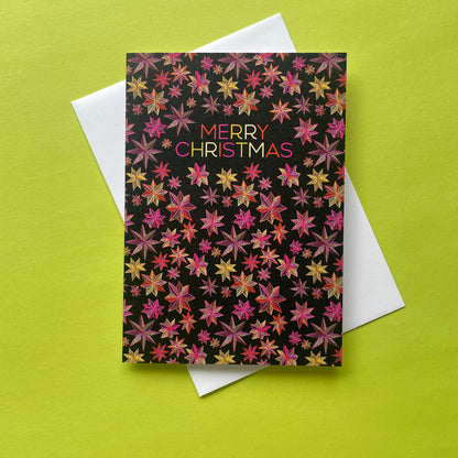 A Christmas greeting card with a black background covered in a dense pattern of pink, red, orange, and gold starburst motifs, with the words 'MERRY CHRISTMAS' printed in the center. The card is shown on top of a white envelope against a bright lime green background.
