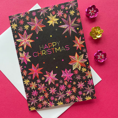 A Christmas greeting card with a black background and colourful star pattern, featuring the words 'HAPPY CHRISTMAS' in pink and gold. The card is styled on top of a white envelope against a bright pink background, with three small metallic flower decorations.