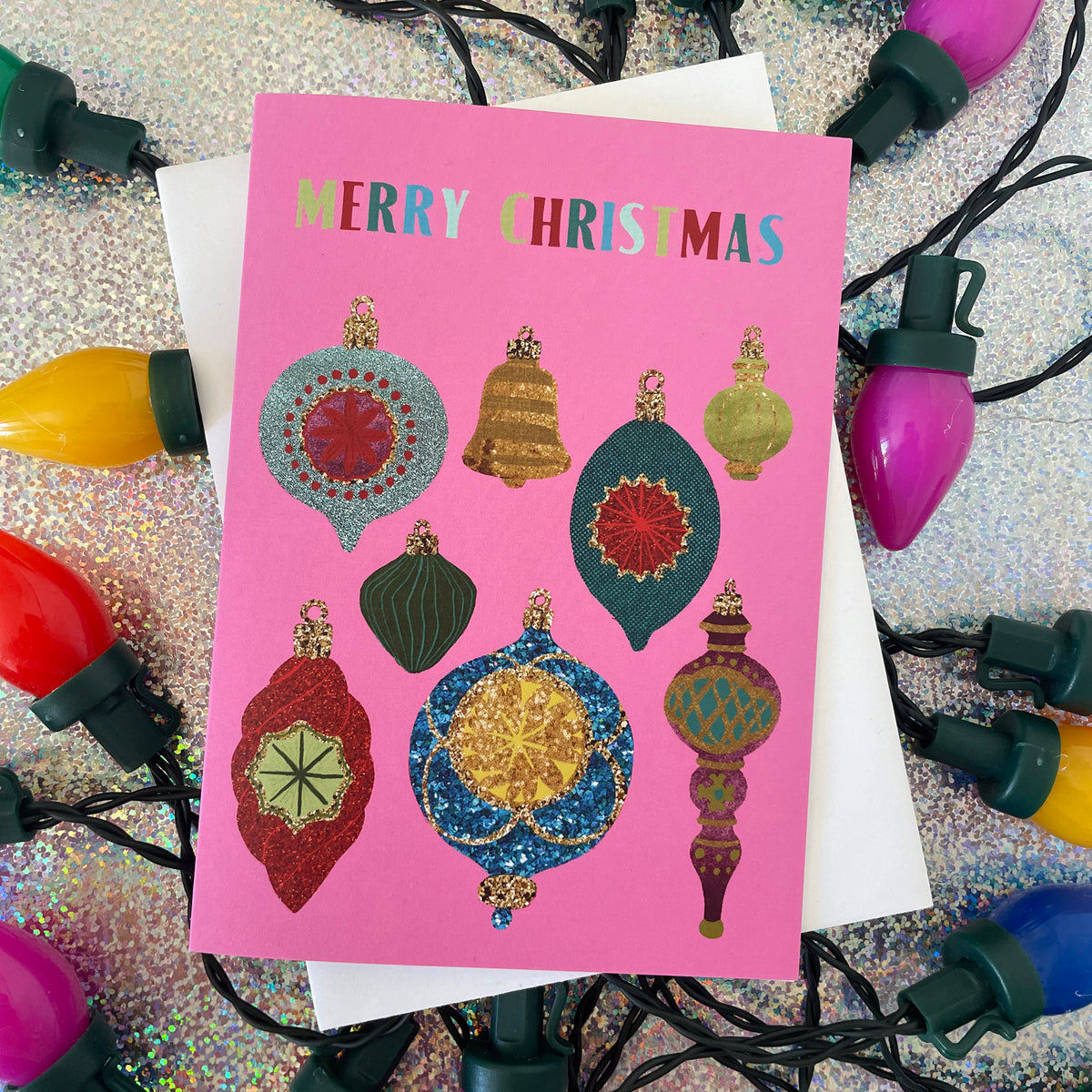 A pink Christmas greeting card with colourful illustrated ornaments and the words “MERRY CHRISTMAS,” photographed on top of white paper and surrounded by multicoloured Christmas lights on a glittery silver background.