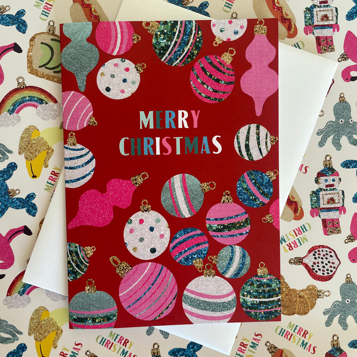A red Christmas greeting card with colourful glittery ornaments and the words 'MERRY CHRISTMAS' in pastel letters, layered over patterned wrapping paper with robots, rainbows, and holiday motifs.