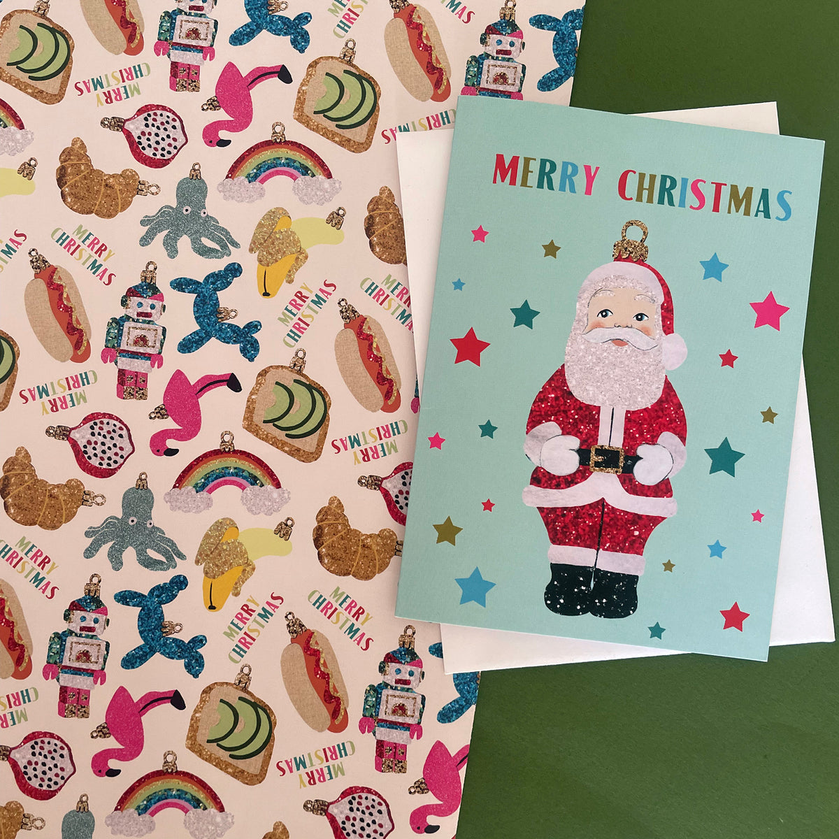 A Christmas greeting card featuring a glittery Santa Claus on a light blue background with colourful stars and the words 'MERRY CHRISTMAS' at the top, layered over a white envelope. The card is placed on top of a pink patterned sheet filled with festive illustrations such as hot dogs, rainbows, snowman ornaments.