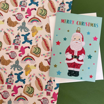 A Christmas greeting card featuring a glittery Santa Claus on a light blue background with colourful stars and the words 'MERRY CHRISTMAS' at the top, layered over a white envelope. The card is placed on top of a pink patterned sheet filled with festive illustrations such as hot dogs, rainbows, snowman ornaments.