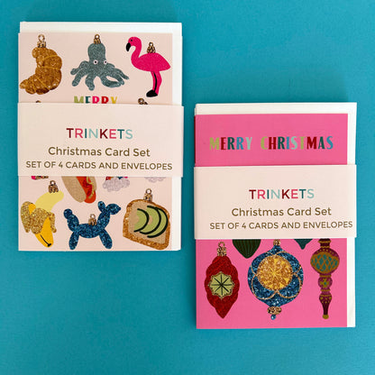 Two Christmas card sets labeled 'TRINKETS Christmas Card Set' arranged on a bright turquoise background. The left set has a cream card with glittery ornament illustrations, and the right set has a pink card with colourful ornament illustrations.