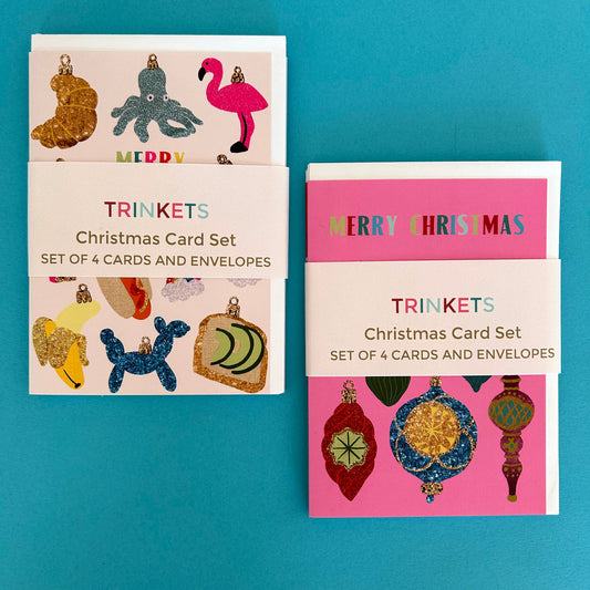 Two Christmas card sets labeled 'TRINKETS Christmas Card Set' arranged on a bright turquoise background. The left set has a cream card with glittery ornament illustrations, and the right set has a pink card with colourful ornament illustrations.