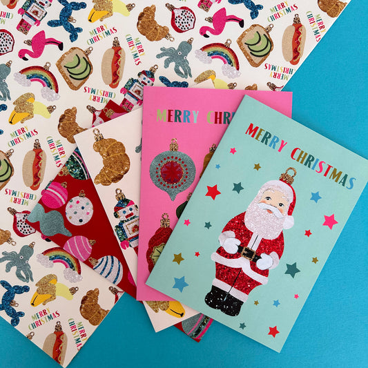Four Christmas greeting cards are arranged on top of glittery wrapping paper. The cards feature festive designs inspired by vintage Christmas ornaments.