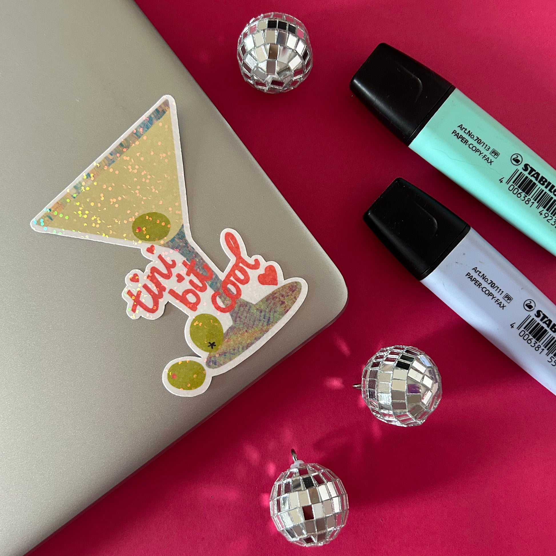 Sticker with 'tini bit cool' text on a notebook with markers and disco balls on a red surface