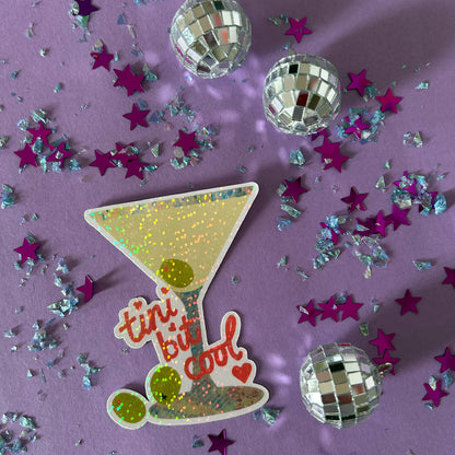 Sticker of a cocktail glass with 'tini bit cool' text on a purple background with disco balls and stars.