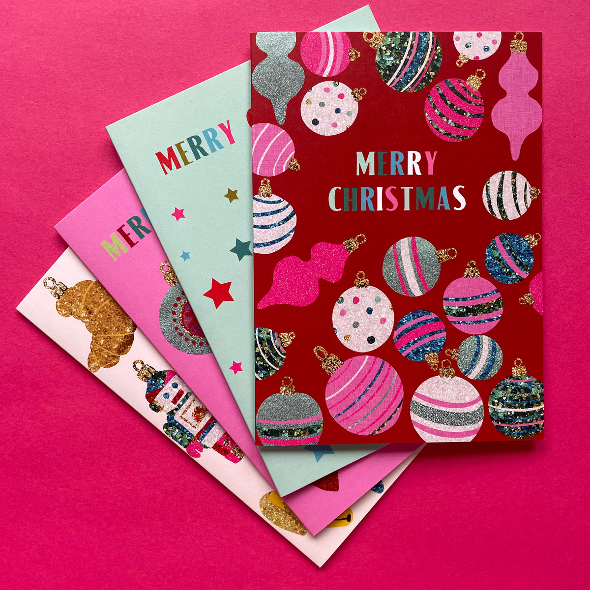 A stack of four Christmas greeting cards fanned out on a bright pink background. The top card is dark red with glittery multicolour Christmas ornaments and the words 'MERRY CHRISTMAS' in white and pink.