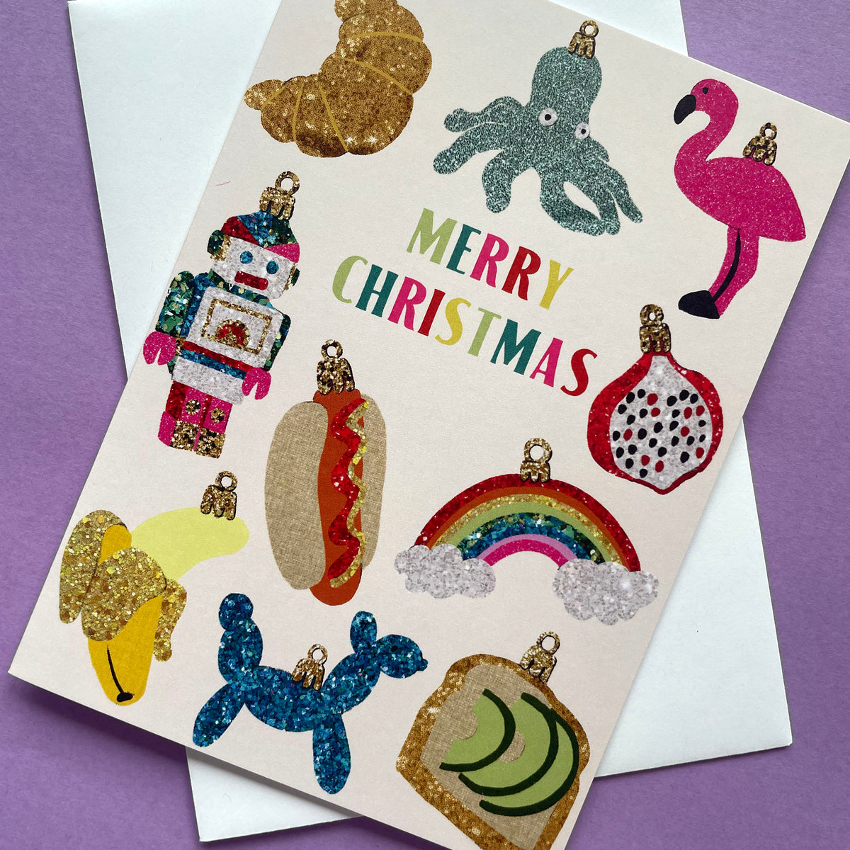 A Christmas greeting card with glittery, colourful ornament illustrations and the words 'MERRY CHRISTMAS' in multicolored letters, shown on top of a white envelope against a purple background.