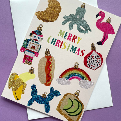 A Christmas greeting card with glittery, colourful ornament illustrations and the words 'MERRY CHRISTMAS' in multicolored letters, shown on top of a white envelope against a purple background.