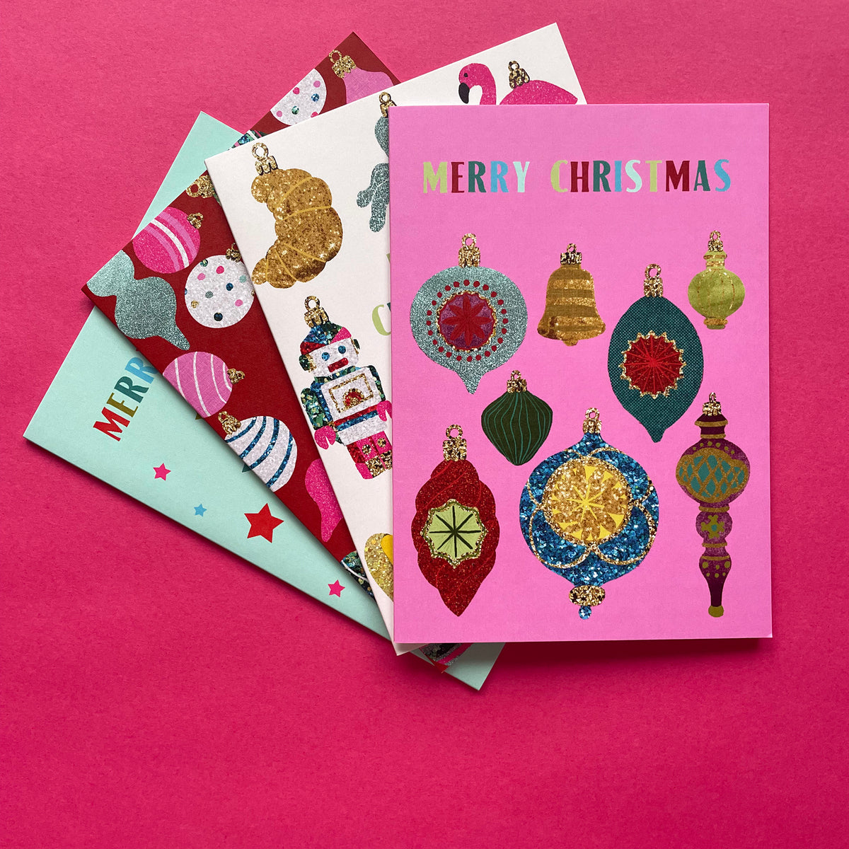 A stack of four Christmas greeting cards fanned out on a bright pink background. The top card is pink with colourful metallic-textured ornaments and the words 'MERRY CHRISTMAS' at the top.