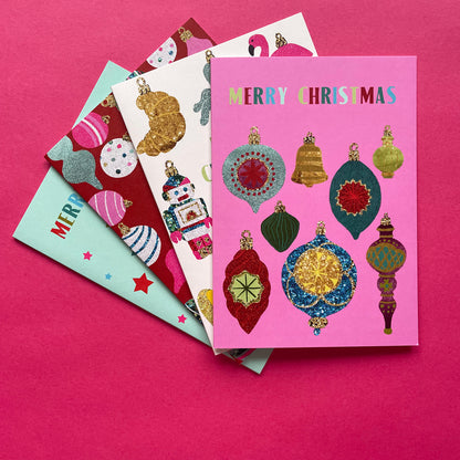 A stack of four Christmas greeting cards fanned out on a bright pink background. The top card is pink with colourful metallic-textured ornaments and the words 'MERRY CHRISTMAS' at the top.