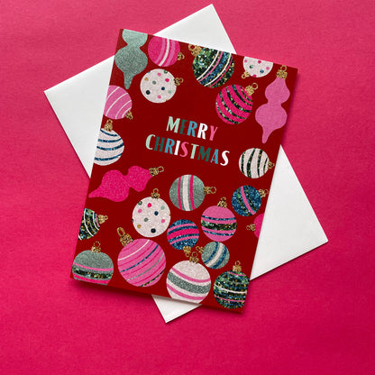 A Christmas greeting card with a red background and colourful illustrated ornaments, placed on top of white envelopes against a bright pink background.