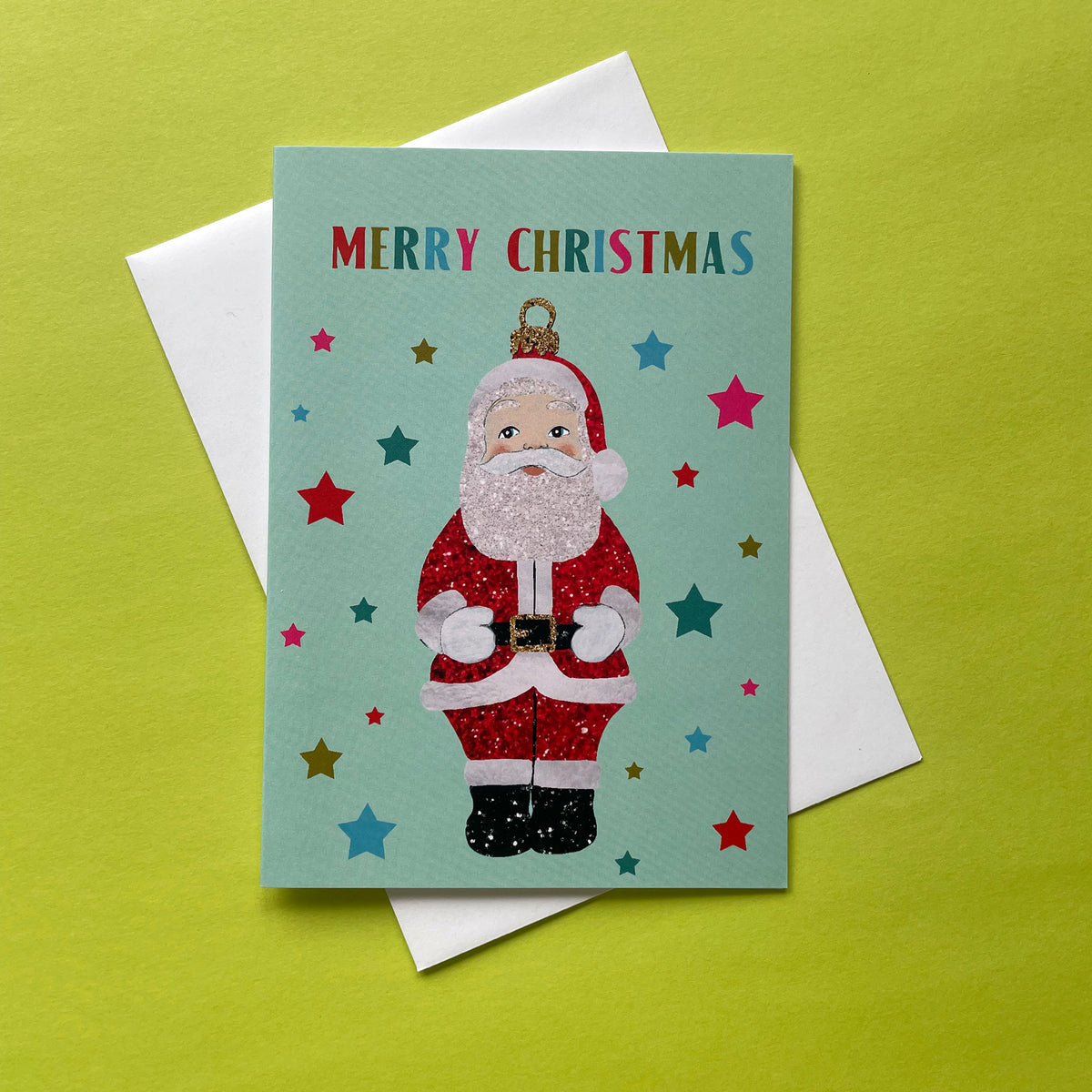 A Christmas greeting card featuring a glittery Santa Claus illustration on a mint green background with colourful stars and the words 'MERRY CHRISTMAS' at the top, shown with a white envelope on a bright yellow-green surface.