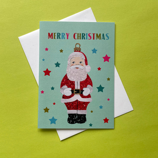 A Christmas greeting card featuring a glittery Santa Claus illustration on a mint green background with colourful stars and the words 'MERRY CHRISTMAS' at the top, shown with a white envelope on a bright yellow-green surface.