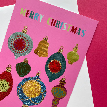 A pink Christmas greeting card with the words 'MERRY CHRISTMAS' in multicoloured letters and several glittery ornament illustrations, shown on top of a white envelope against a magenta background.