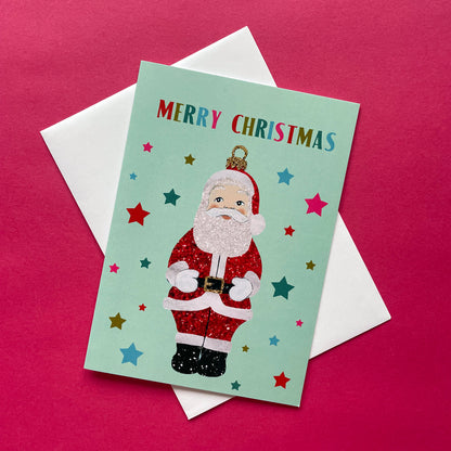 A Christmas greeting card featuring a glittery Santa Claus on a mint green background with colourful stars and the words 'MERRY CHRISTMAS' at the top, shown with a white envelope on a bright pink background.