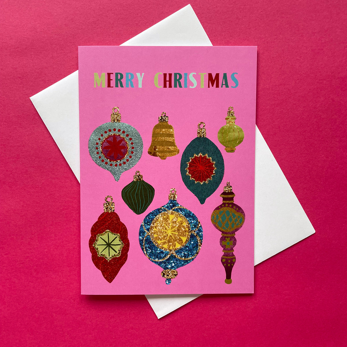 A pink Christmas greeting card with colourful ornament illustrations and the words 'MERRY CHRISTMAS' at the top, placed on top of a white envelope against a bright pink background.