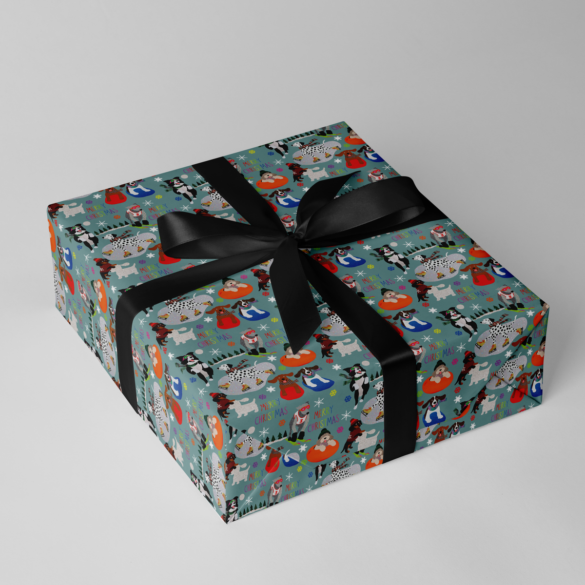 A square gift box wrapped in teal wrapping paper with a colourful dog pattern and tied with a wide black ribbon bow, photographed on a light grey studio background.