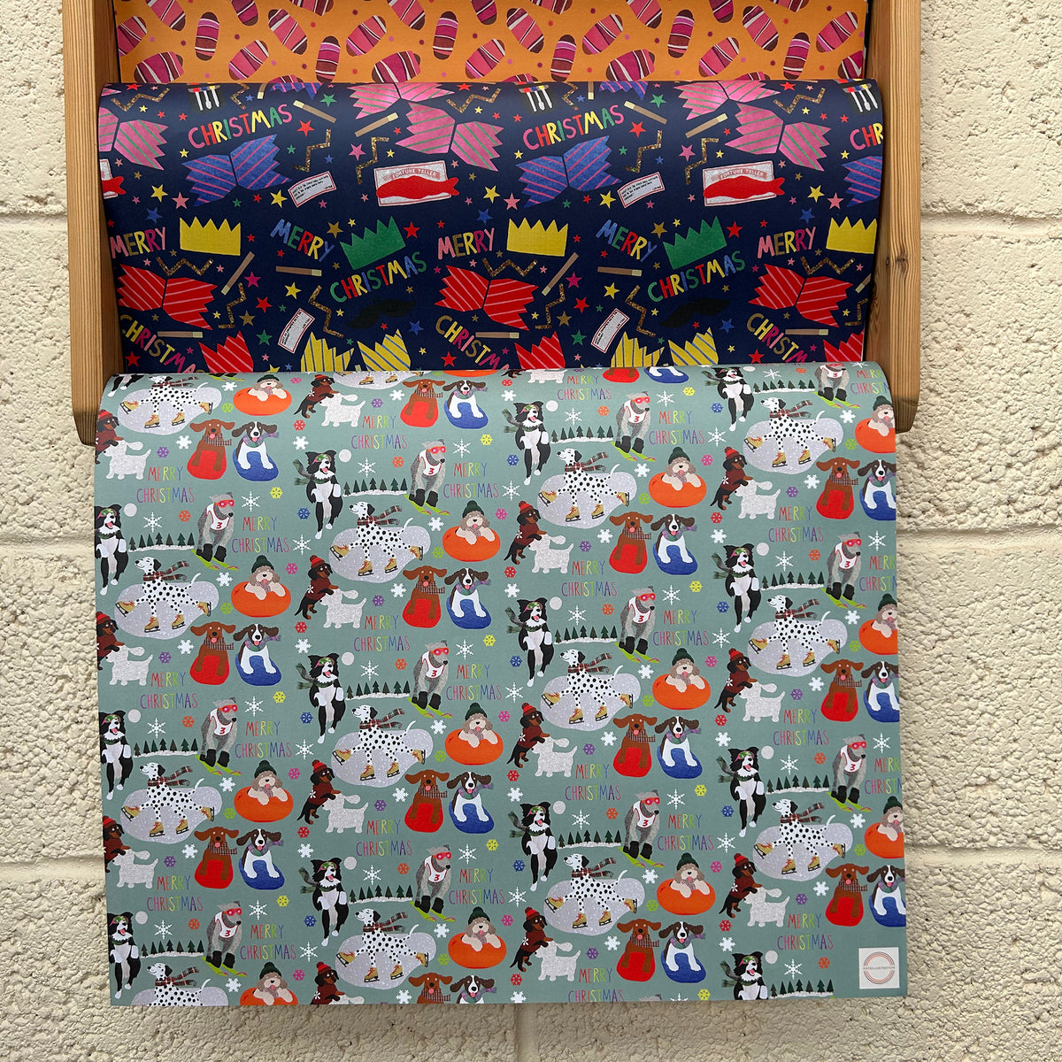 Three rolls of brightly coloured Christmas-themed wrapping paper hanging on a wooden hanger against a light beige brick wall. 