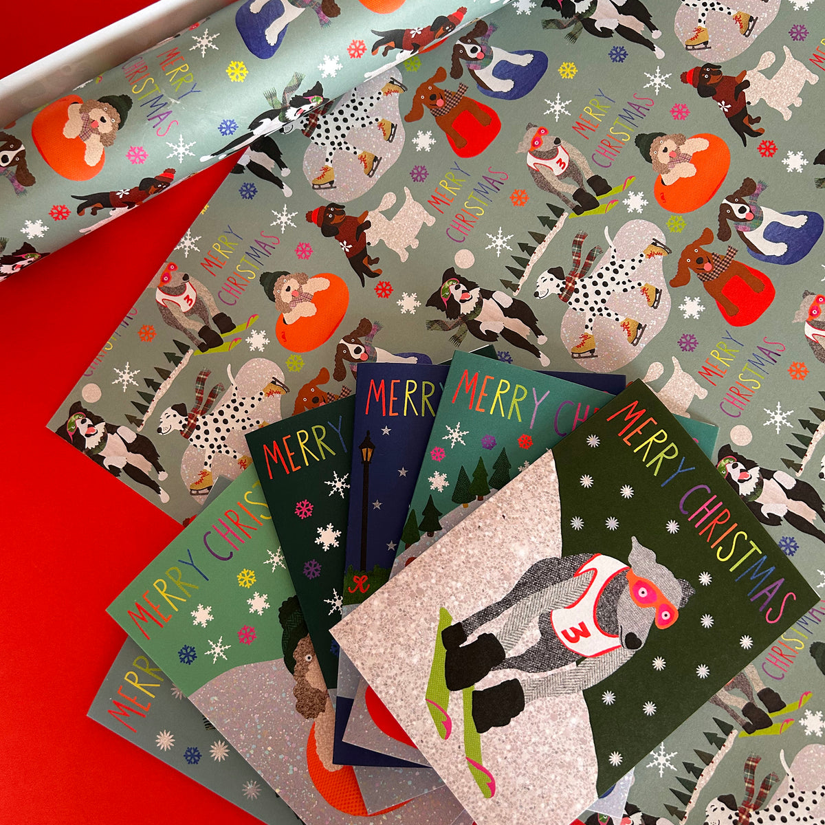 A collection of Christmas-themed gift wrap and greeting cards featuring a whimsical dog illustration pattern and a snowy ski dog design, arranged on a bright red surface.