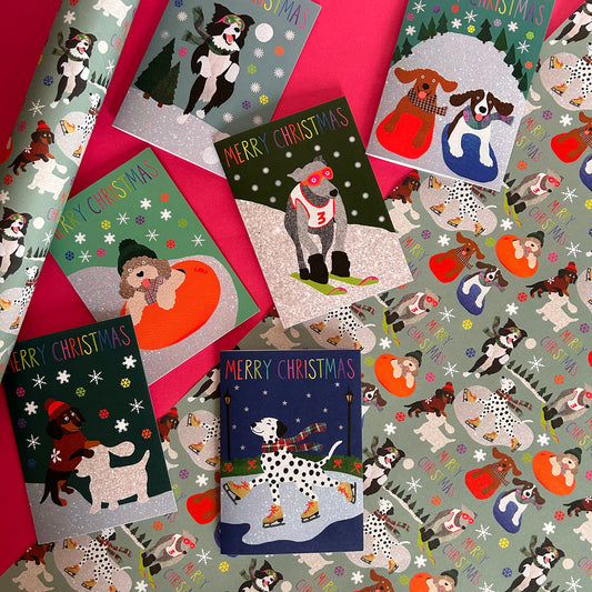 A flat lay of Christmas gift cards and wrapping paper featuring illustrated dogs in winter scenes, including a greyhound skiing, a collie catching snowballs, and a dalmatian at the skate rink. The cards say Merry Christmas and are arranged on a bright pink background with matching patterned wrapping paper visible around the edges.