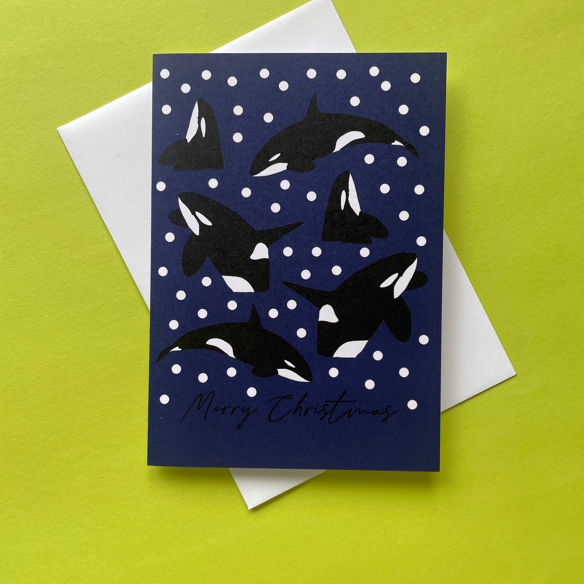 A navy blue Christmas greeting card featuring black and white orca illustrations on a white snowball background, with 'Merry Christmas' written in black script. A white envelope is partially visible behind the card, which is placed on a bright yellow-green surface.