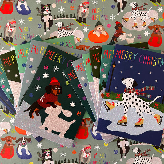 A fan of Christmas greeting cards featuring illustrated dogs in winter holiday scenes, including a Dalmatian ice skating and a dachshund building a snow dog, with 'MERRY CHRISTMAS' text on a patterned blue grey background.