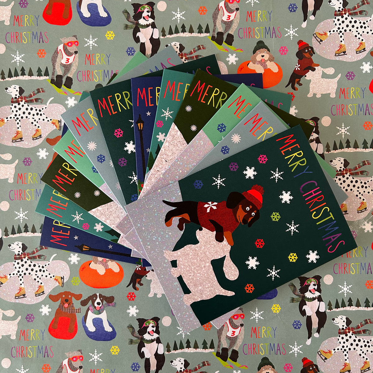 A fan of Christmas greeting cards featuring illustrated dogs and the text 'MERRY CHRISTMAS' on a grey patterned background with matching dog-themed wrap.