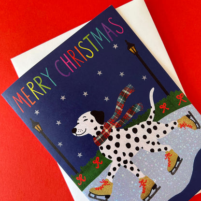 A Christmas greeting card featuring a Dalmatian dog ice skating at night under a dark blue sky, with the words 'MERRY CHRISTMAS' in rainbow letters. The card is shown at an angle on top of a white envelope against a solid red background.