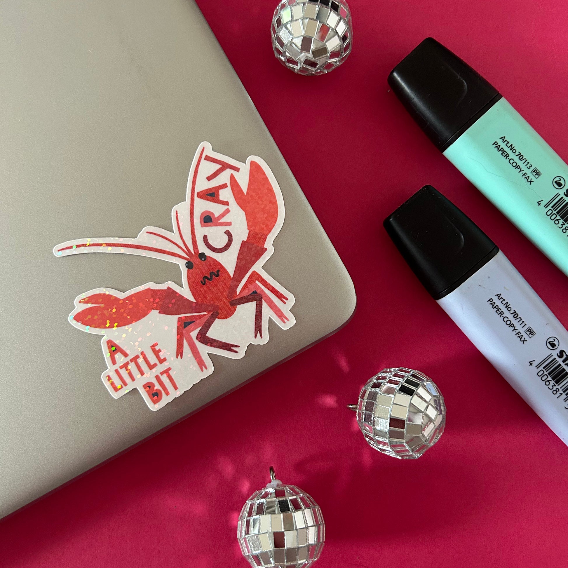 Sticker of a red crayfish with text on a laptop, surrounded by disco balls and markers.